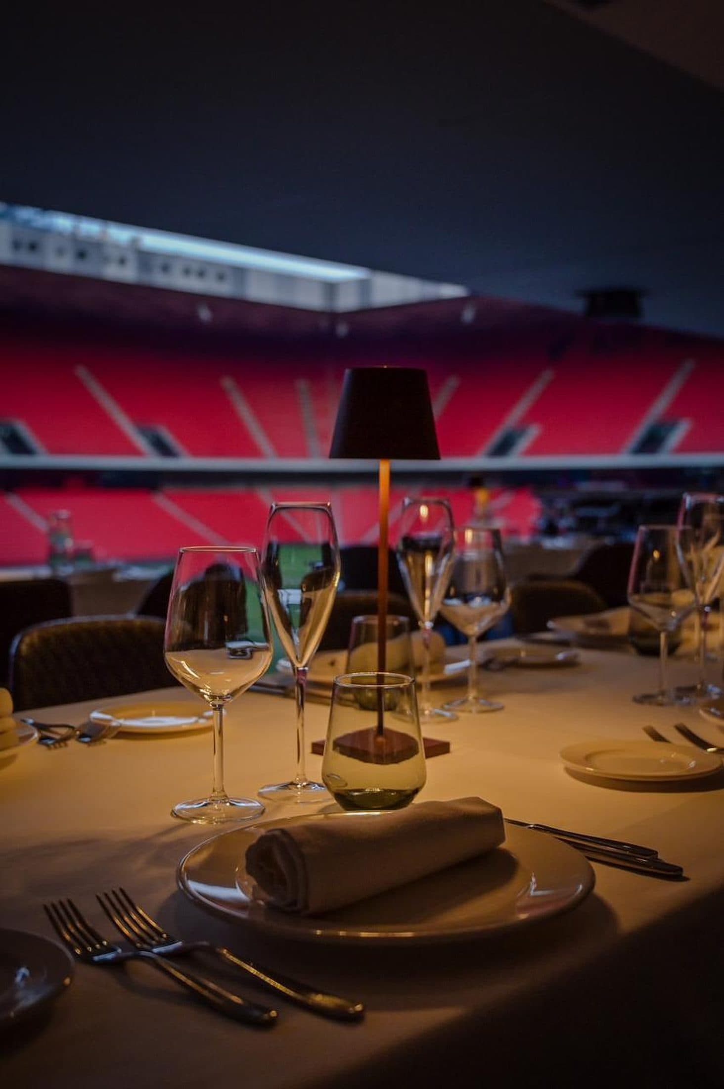 A copper table lamp glowing at a table set for dinner, the empty red stadium seats and a glimpse of the pitch behind