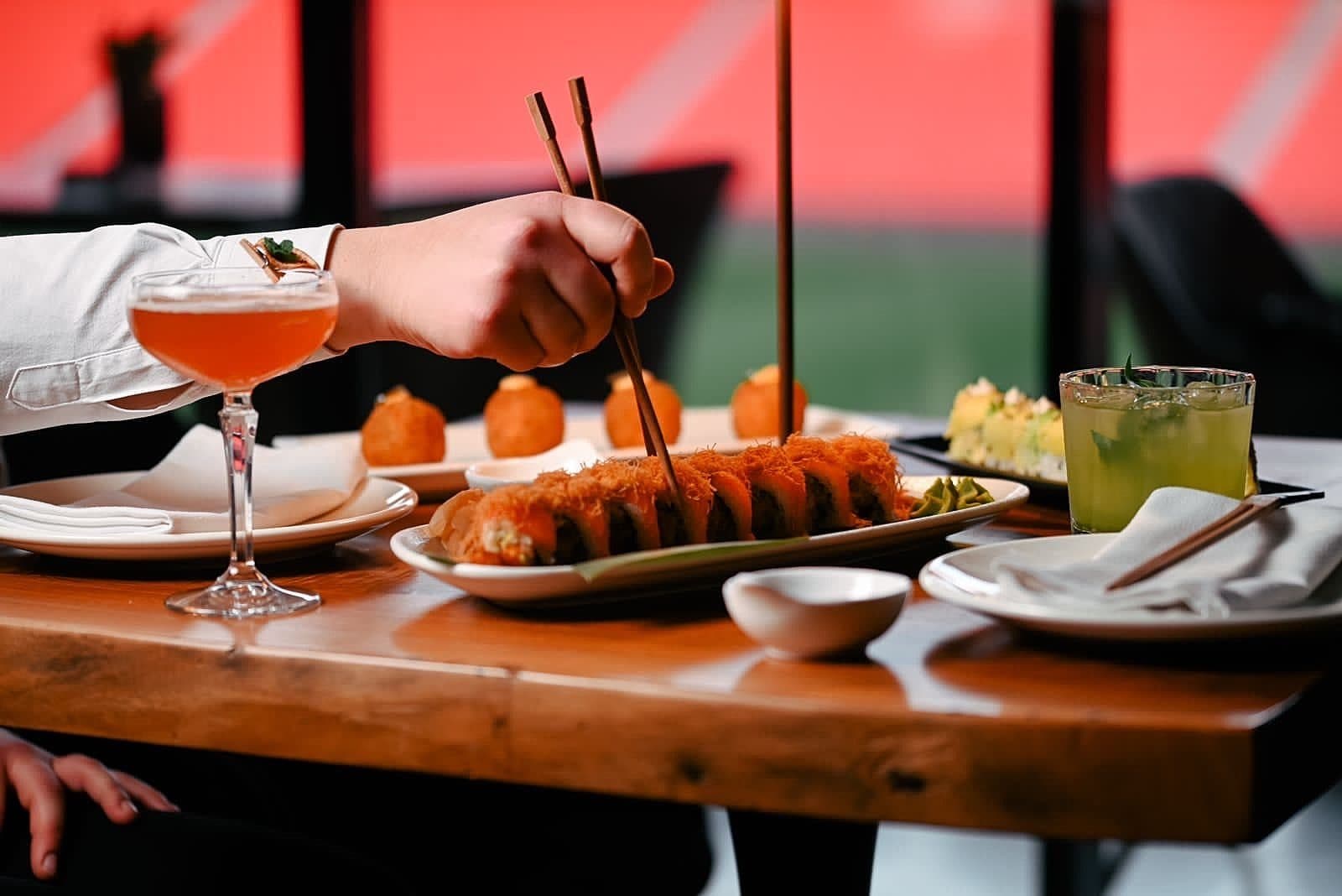 Hand using chopsticks to lift a sushi roll from a platter on the bar table, the stadium pitch visible behind