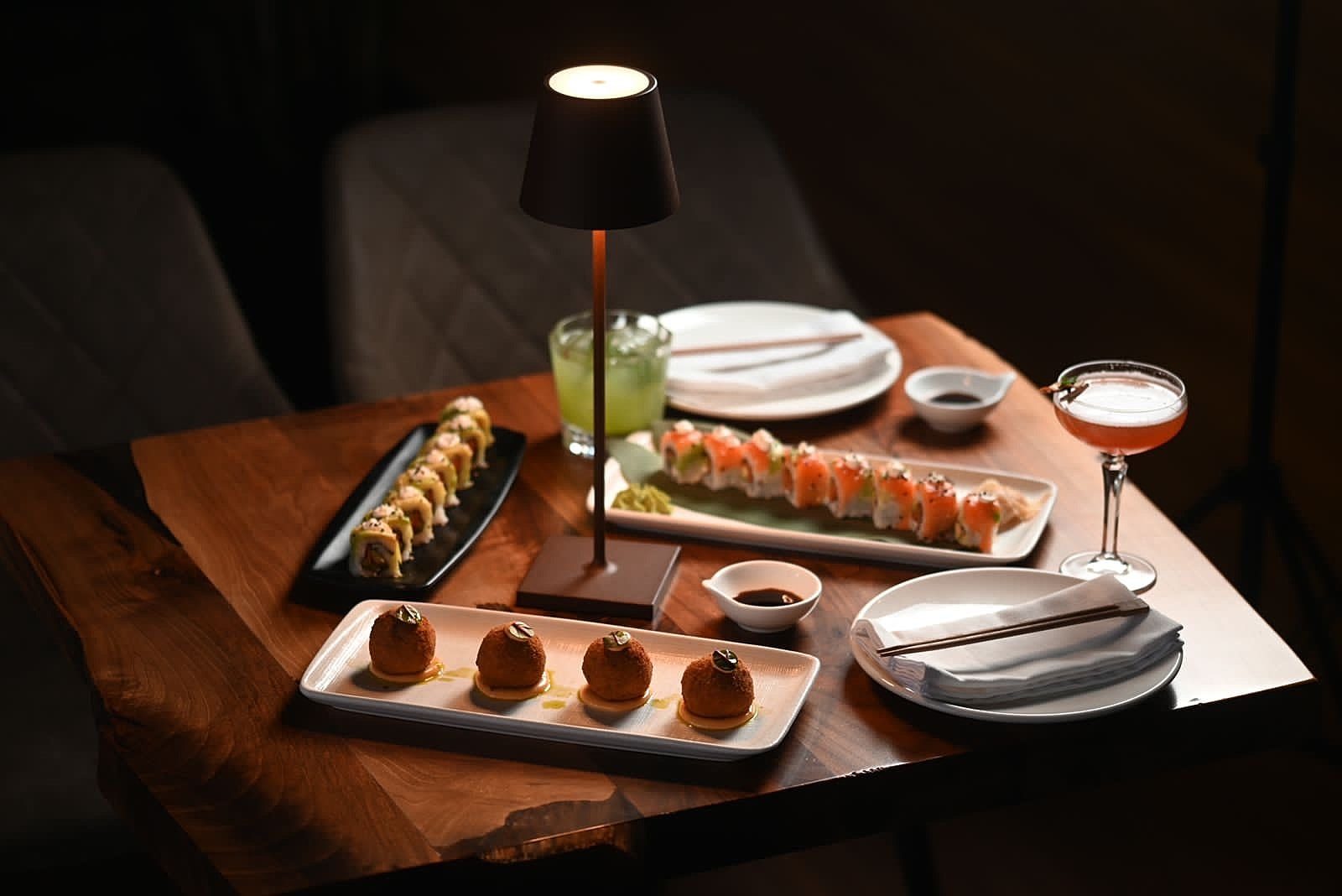 A copper table lamp beside a sushi platter, cocktail and small dessert bites set on the bar table