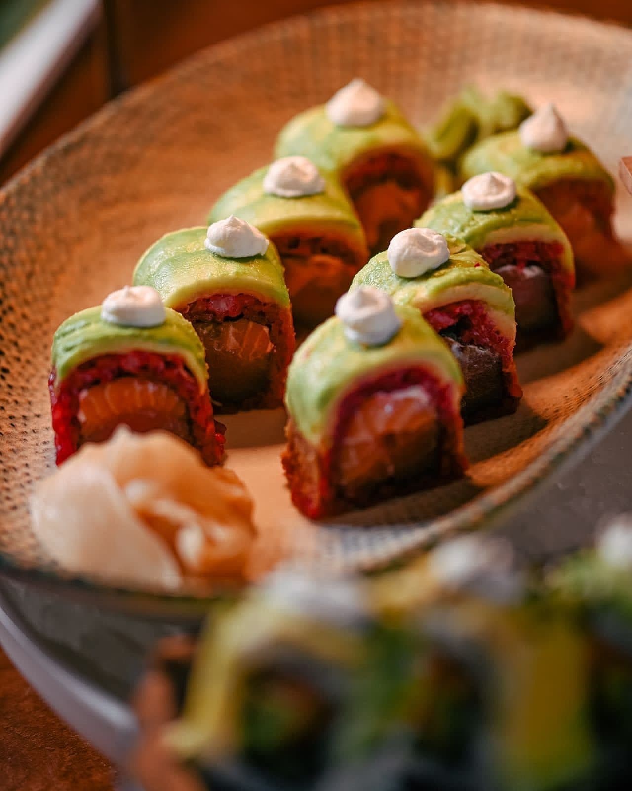 Avocado-wrapped sushi rolls with sauce dollops and pickled ginger, plated on a rattan tray
