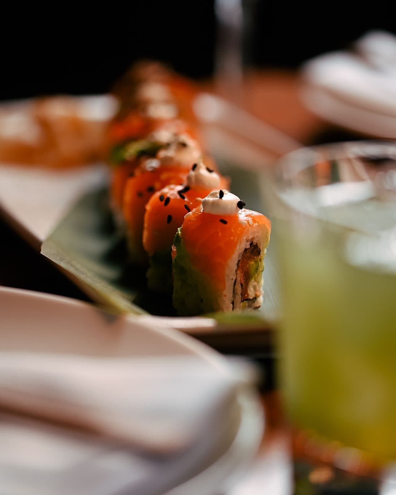 A salmon sushi roll platter on the bar table with a green cocktail blurred in the foreground